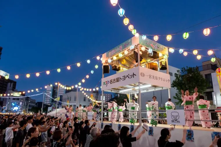 夜の一宮七夕まつりで提灯が連なる盆踊りやぐらの上、浴衣姿の踊り手たちが両手を上げて盆踊りを披露する様子。やぐらの側面には協賛企業の看板が並び、周囲を大勢の観客が取り囲んでいる夜間の賑わい。