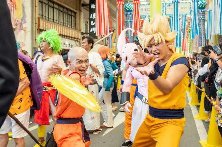 一宮七夕まつりのコスプレパレードで人気アニメキャラクターに仮装した参加者たち。オレンジの道着姿のドラゴンボール孫悟空風コスプレイヤーが技を繰り出すポーズを取り、隣で緑髪や坊主頭のキャラクターに扮した参加者も表情豊かに演技している。背景には極彩色の七夕飾りが下がる。