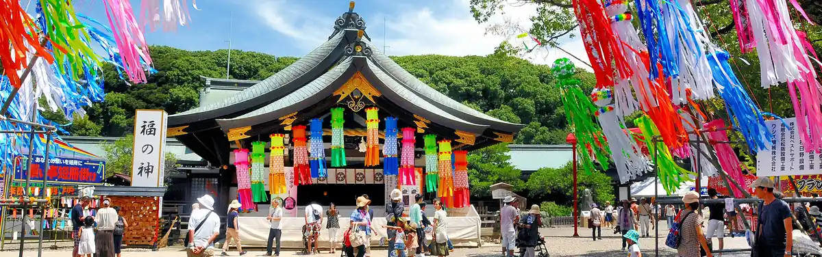 一宮七夕まつり公式サイトのメインビジュアルにも使われている、真清田神社境内の七夕飾りの様子。極彩色の吹き流しと短冊が並び、浴衣や帽子姿の来場者たちが行き交う晴天の日中の光景。左に「福の神」の看板と願い事短冊掛けが見える。