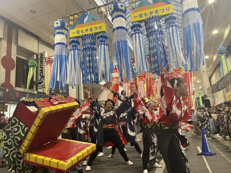 ワッショーいちのみやで本町商店街を練り歩く連の様子。赤い法被姿の踊り手たちが山車を先頭に、色鮮やかな衣装で通りいっぱいに広がって踊っている。頭上のアーケードには七夕飾り、沿道には大勢の観客が集まっている。