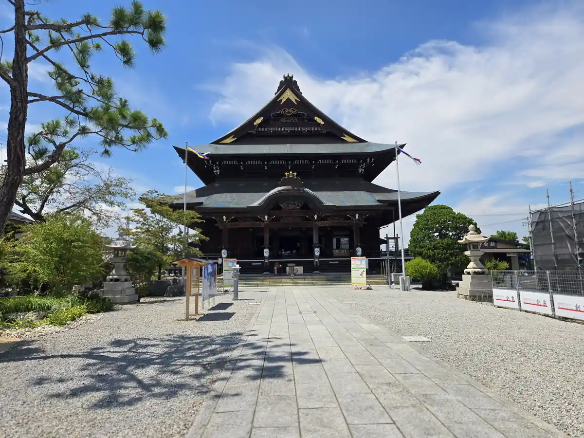松の木の奥に善光寺東海別院の本堂が見える境内の参道の写真。石畳の参道の両脇に石灯籠と案内看板が並び、本堂前には多色の幟が掲げられている。右手には工事中の隣接建物も見え、晴天で参拝者の姿はない。