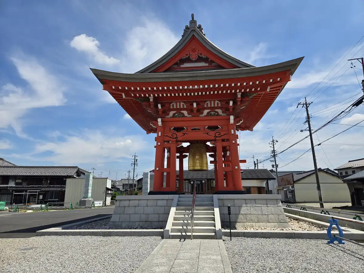 善光寺東海別院の朱塗りの鐘楼を正面から見上げるように撮影した写真。大きく反った軒の下に金色の梵鐘が吊るされ、石積みの基壇から石段と手すりが鐘楼へ続く。奥には住宅や電柱が写り込み、晴天の空が広がる。