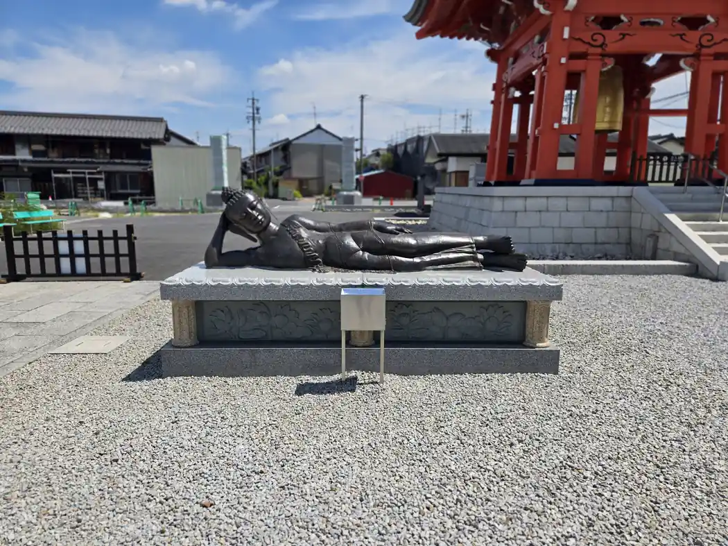 石造りの台座に横たわる寝釈迦像の写真。右手で頭を支えながら横になった姿で、台座の側面には蓮の花の彫刻が施されている。背景には朱塗りの鐘楼の屋根と金色の梵鐘、遠くには住宅街が写る。