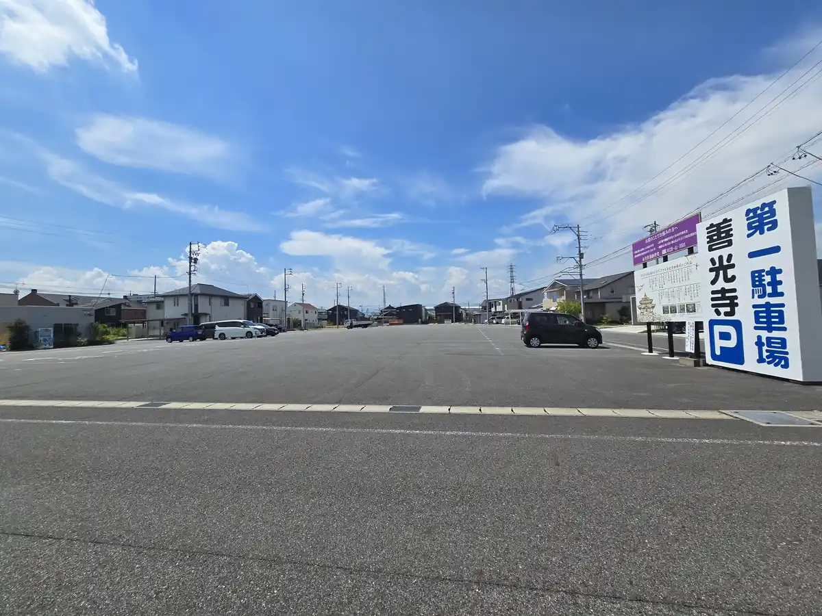 善光寺東海別院の第一駐車場の写真。広いアスファルトの駐車スペースに数台の車が停まり、右手に「第一駐車場 善光寺」と書かれた大きな案内看板が立つ。晴天で駐車場の奥まで広く見渡せる様子が写る。