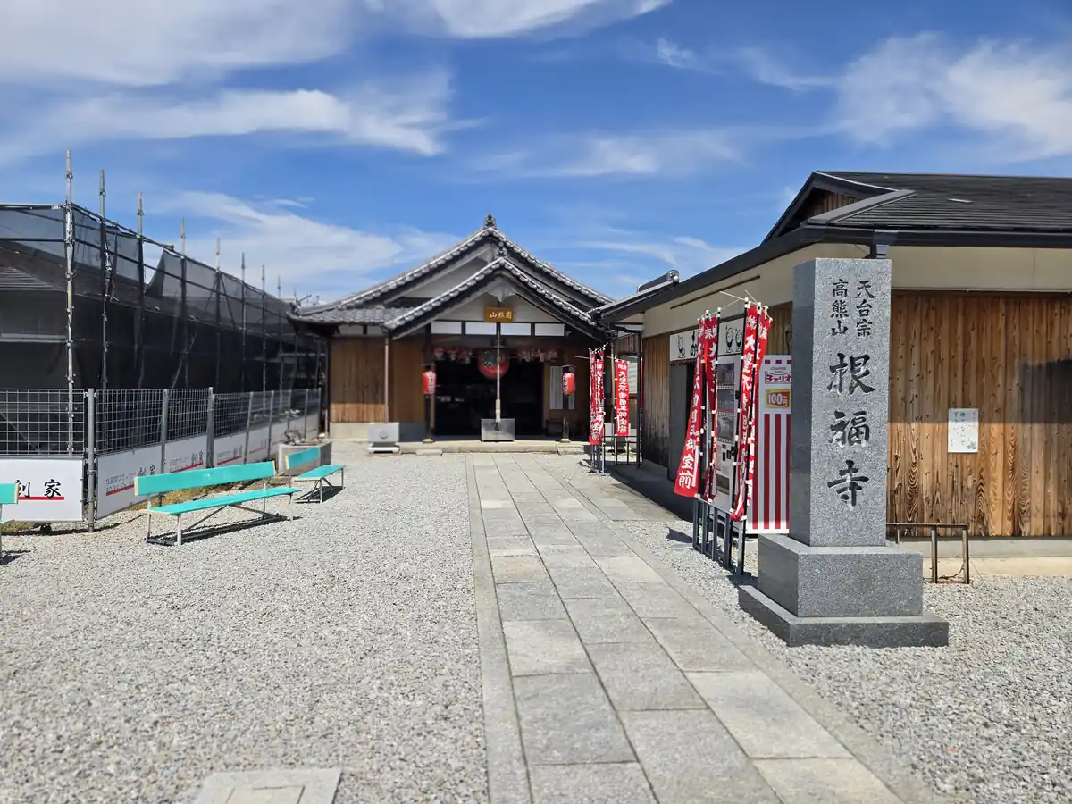 工事用フェンスの奥に見える根福寺の写真。「天台宗 高熊山 根福寺」と刻まれた石碑が立ち、山門には赤い幟が何本も掲げられている。左手には足場を組んだ工事中の建物があり、右手前にはベンチが置かれている。