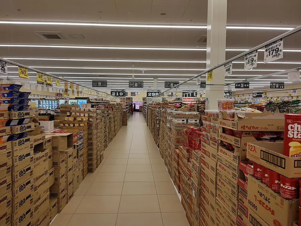 ラ・ムー稲沢店のお菓子売場の通路を写した様子。両側の棚にカップ麺やスナック菓子が開封したダンボール箱に入ったまま積み重ねて陳列され、黄色い価格POPが多数吊り下げられ、ベージュのタイル床がまっすぐ奥まで続いている。