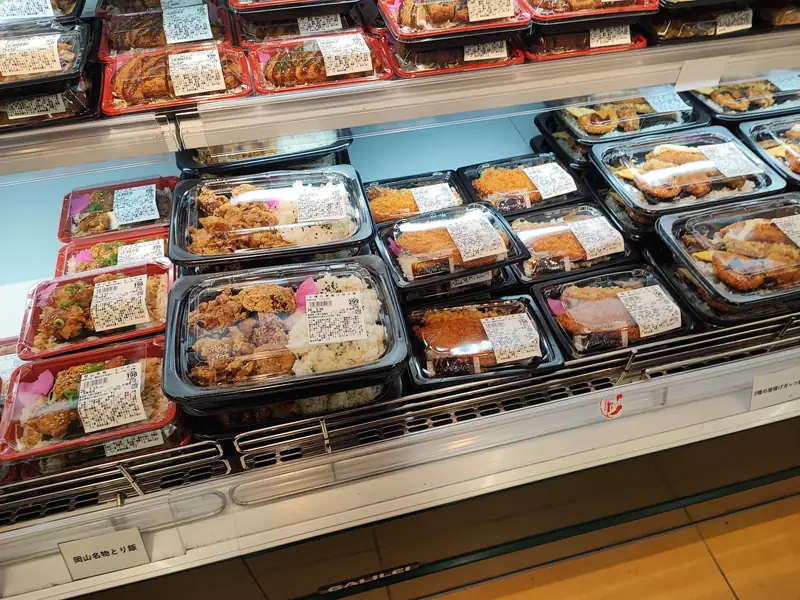 ラ・ムー稲沢店の惣菜コーナーの弁当売り場を斜め上から写した様子。唐揚げ弁当、唐揚げとカツを盛ったボリューム満点の丼、岡山名物とり飯など、赤や黒の容器に入った弁当が多数陳列され、198円から299円ほどの値札が付いている。