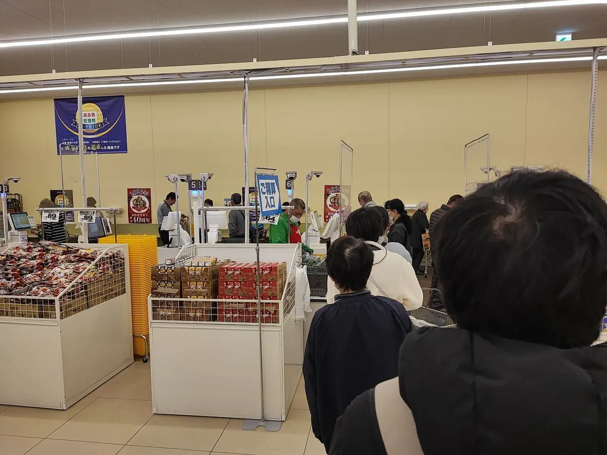 ラ・ムー稲沢店のレジカウンター前に買い物客が列をつくる店内の様子。黄色い買い物カゴが積み上げられ、奥には緑色のエプロンをつけた店員や精算機とみられる機械が並んでいる。天井の照明が店内全体を明るく照らしている。