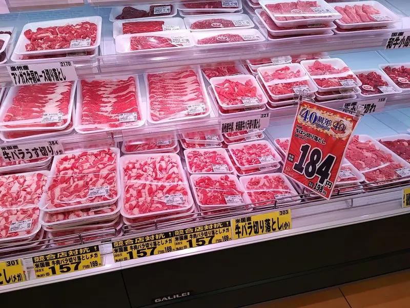 ラ・ムー稲沢店の精肉売り場の冷蔵ケースを写した様子。牛バラ肉やロース肉など様々な部位の牛肉パックが整然と並び、赤い40周年記念POPに牛ばら切り落とし100gあたり184円と大きく表示され、赤身と脂身のコントラストが鮮やかに写っている。