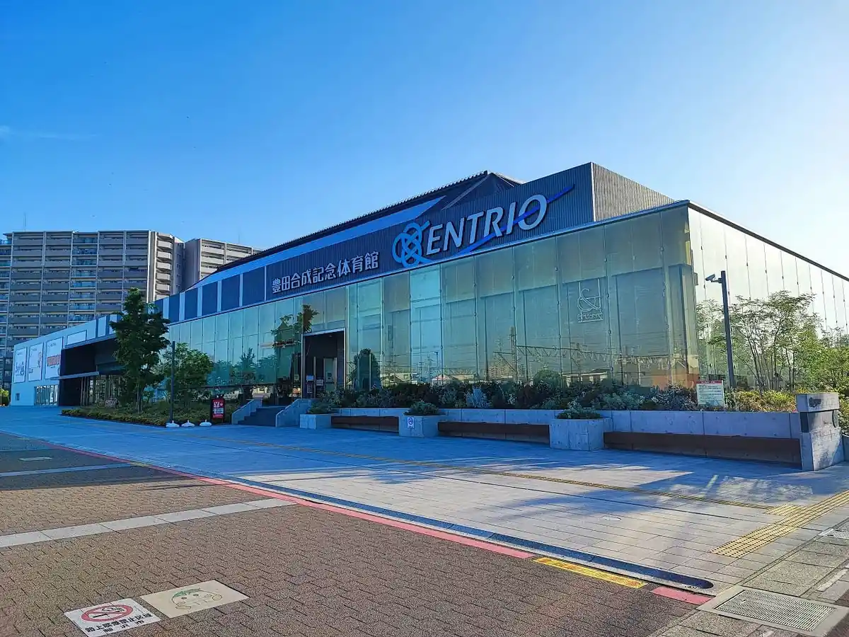 「豊田合成記念体育館ENTRIO」の文字が入った建物外観。ガラス張りの大きな体育館で、手前に植栽と屋外ベンチが並ぶ広場がある。青空に映える建物正面の様子。