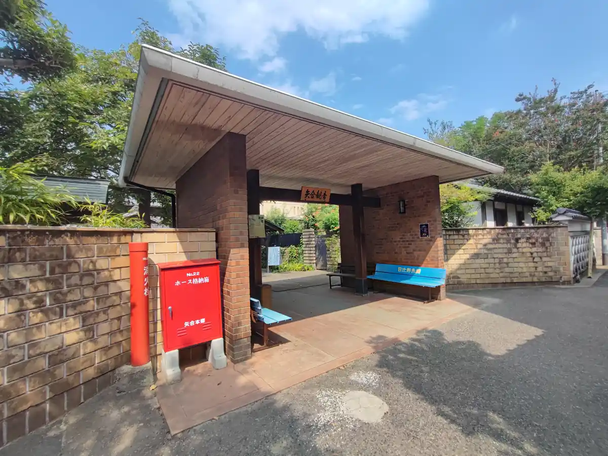 矢合観音の入口の門。レンガ造りの門柱に木の屋根がかかり、「矢合観音」と書かれた木製看板が掲げられている。門の下にはベンチが置かれ、参拝者が一息つけるようになっている。