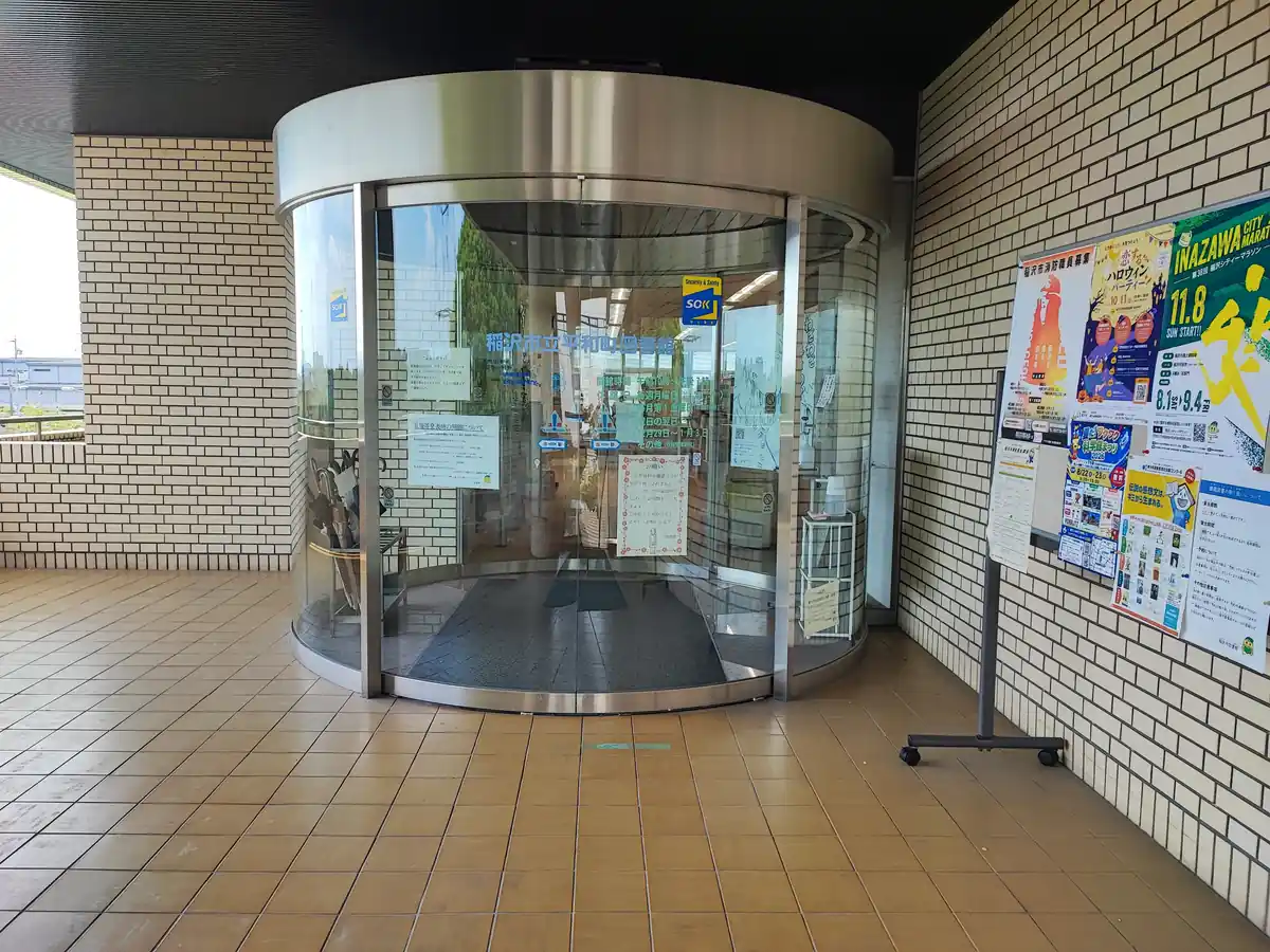 平和町図書館の入口にある半円形のガラス自動ドア正面。ガラス面に「稲沢市立平和町図書館」の文字と開館時間の案内が貼られ、扉の右手の掲示板にはハロウィンイベントや稲沢シティマラソンなど地域のお知らせポスターが並んで掲示されている。