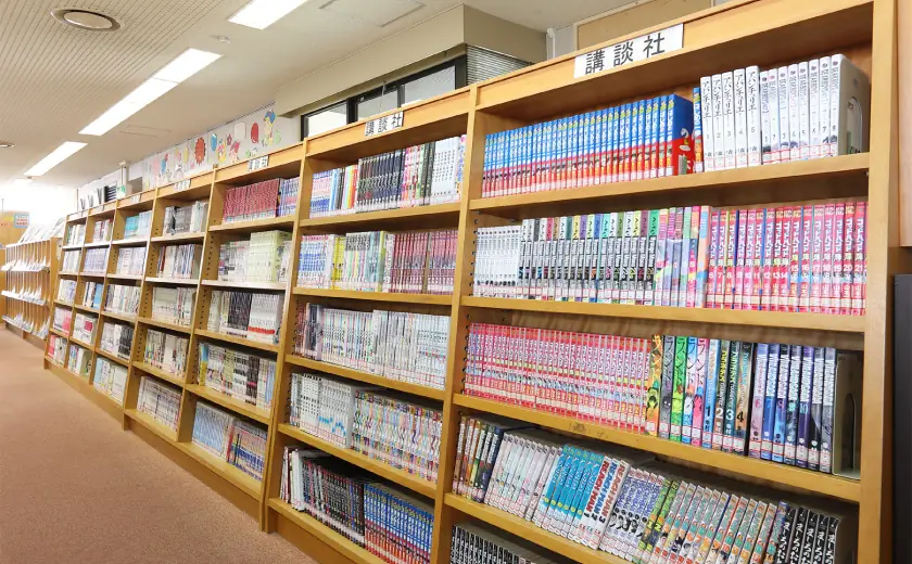 平和町図書館の館内、木製の書棚に単行本サイズのコミックが隙間なく並ぶ書架。背表紙のタイトルが多数見え、漫画コーナーの充実ぶりが伝わる。