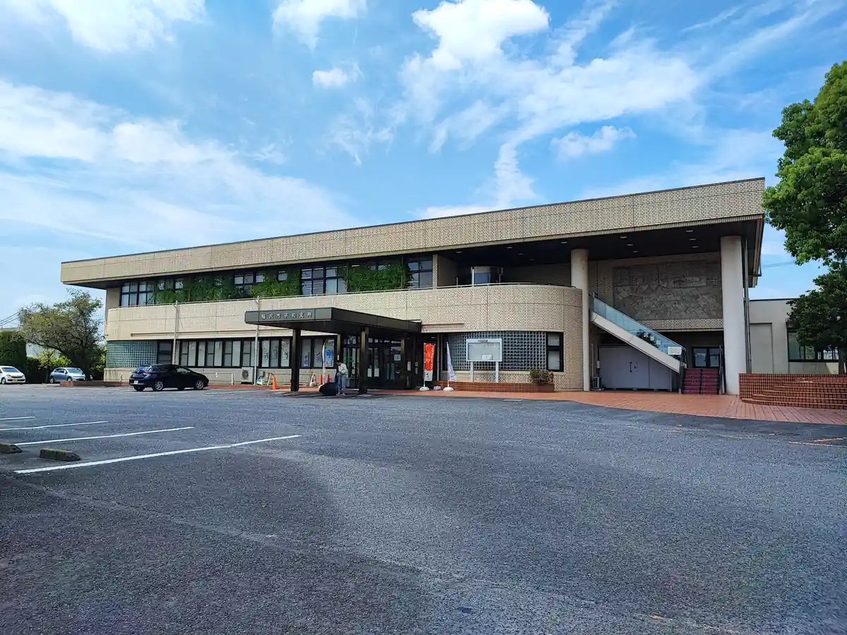 稲沢市平和支所の建物全景。手前に広い駐車場が広がり、ベージュのタイル張り2階建ての建物正面に「稲沢市平和支所」の看板が見える。建物右側には2階にある平和町図書館へ直接上がれる屋外階段が設置されている。