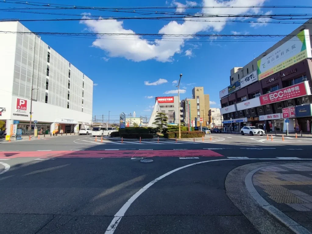 名鉄国府宮駅前のロータリー。白い立体駐車場ビルを背に、駅名の案内看板と円錐形の記念モニュメントが立ち、タクシー乗り場や周辺の商店が並ぶ交差点の様子。