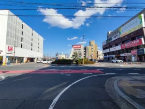 名鉄国府宮駅前のロータリー。白い立体駐車場ビルを背に、駅名の案内看板と円錐形の記念モニュメントが立ち、タクシー乗り場や周辺の商店が並ぶ交差点の様子。