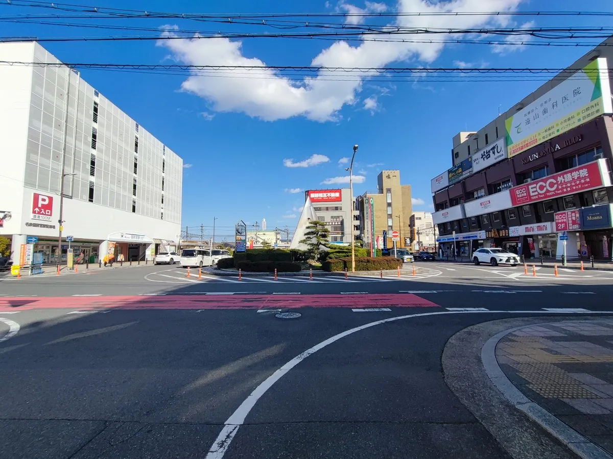 名鉄国府宮駅前のロータリー。白い立体駐車場ビルを背に、駅名の案内看板と円錐形の記念モニュメントが立ち、タクシー乗り場や周辺の商店が並ぶ交差点の様子。