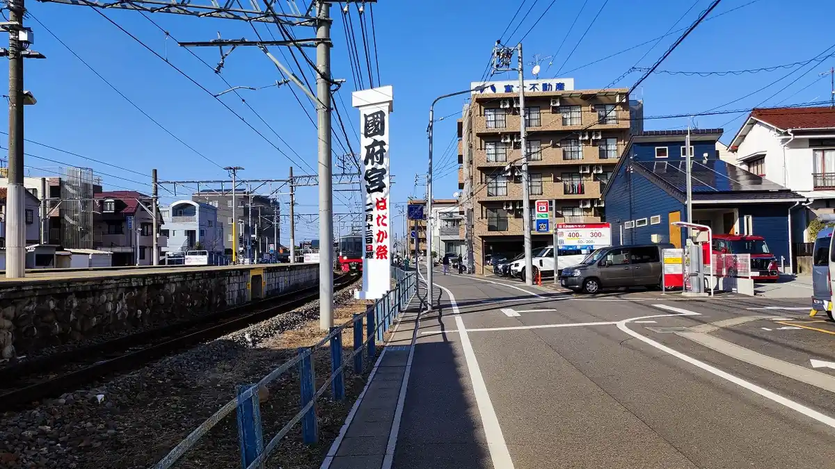 線路沿いの道に立つ「國府宮 はだか祭 二月十三日」と書かれた白い案内塔。奥に茶色いマンションと不動産店の看板、コインパーキングの料金表示が見える駅前通りの風景。