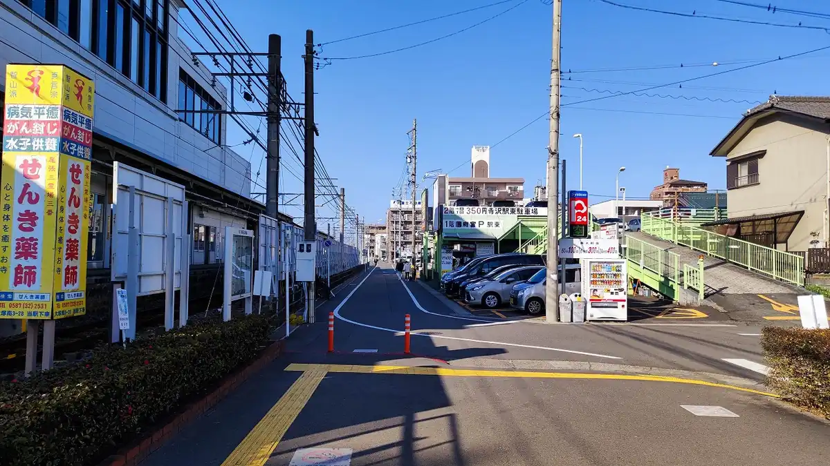 線路沿いの通路にある「寺沢駅東駐車場」「国府宮P 駅東」のコインパーキング案内看板と、隣に設置された飲料の自動販売機。奥に緑色の歩道橋が見える。