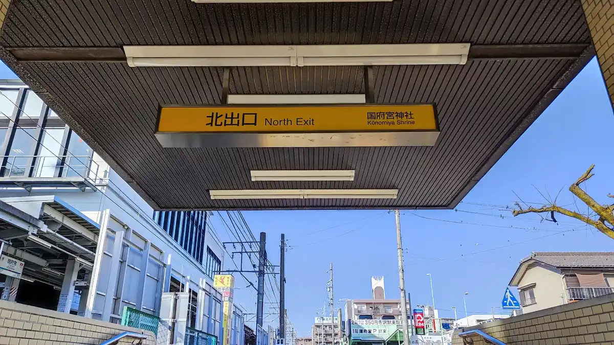 駅の屋根下に掲げられた「北出口 North Exit 国府宮神社 Kōnomiya Shrine」の案内サイン。サインの奥に出口へ続く階段と町並みが見える。
