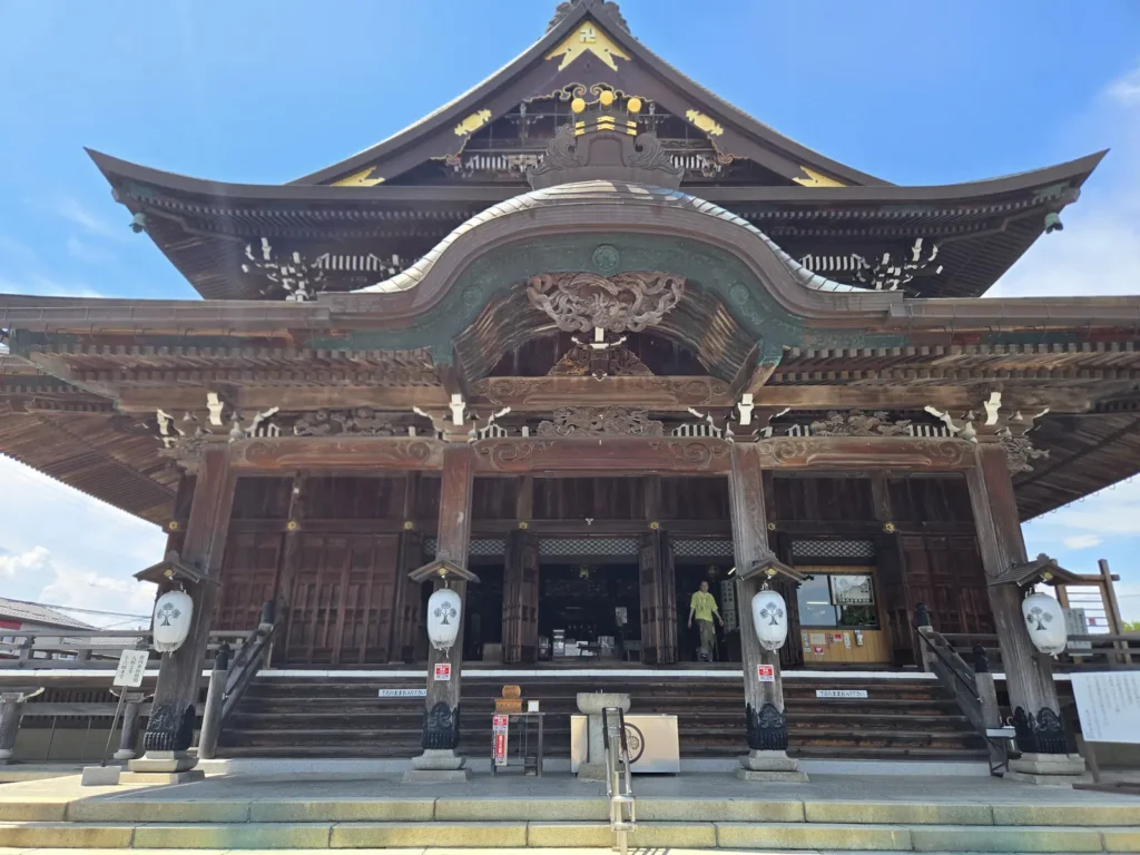 善光寺東海別院の本堂を石段の下から見上げるように撮影した写真。唐破風の大きな向拝と入母屋造りの屋根、金色の飾り金具、左右に下がる白い提灯、両脇に並ぶ石灯籠が写る。晴天の下、本堂の中から出てくる参拝者の姿も小さく写り込んでいる。