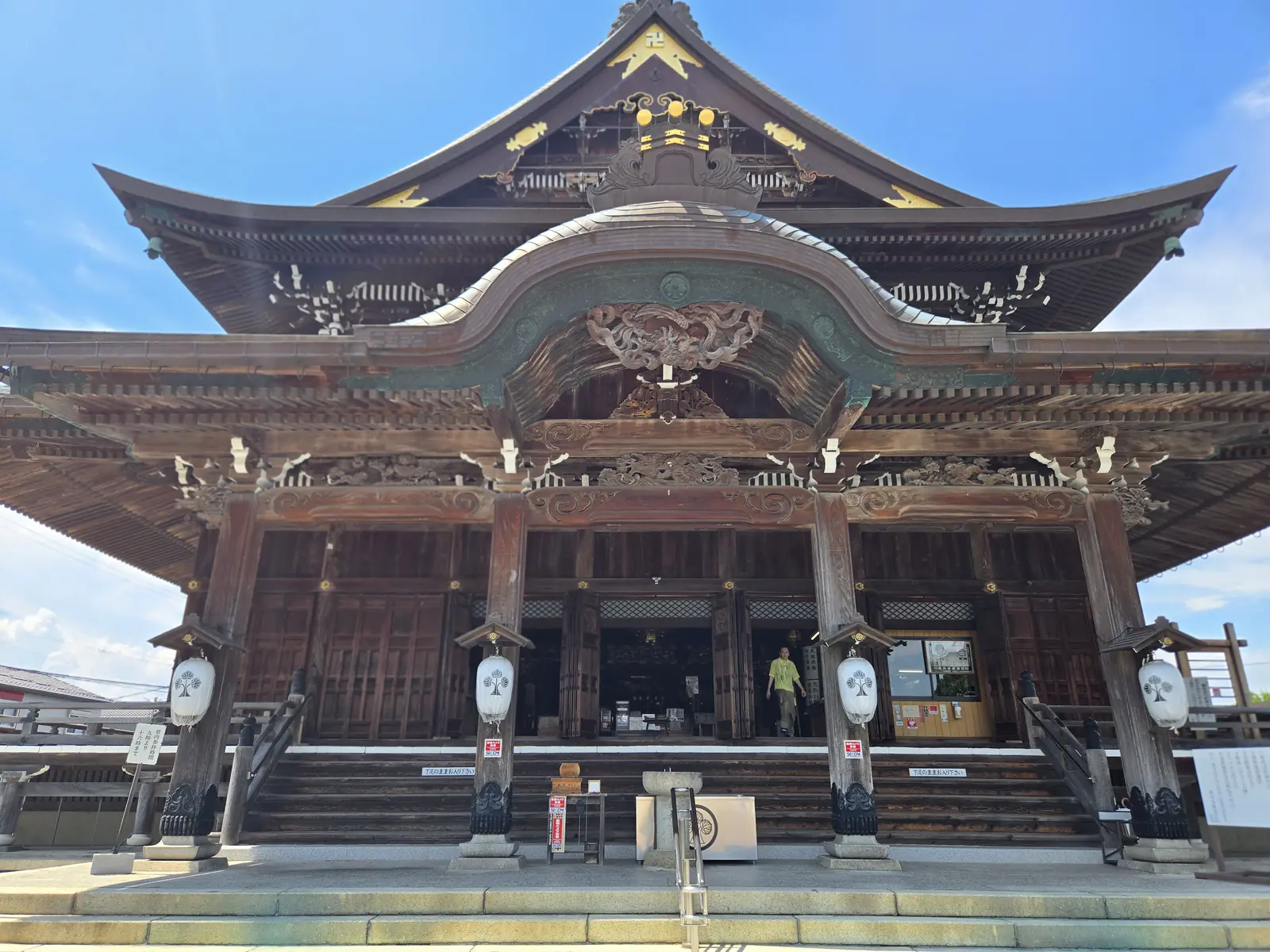 善光寺東海別院の本堂を石段の下から見上げるように撮影した写真。唐破風の大きな向拝と入母屋造りの屋根、金色の飾り金具、左右に下がる白い提灯、両脇に並ぶ石灯籠が写る。晴天の下、本堂の中から出てくる参拝者の姿も小さく写り込んでいる。