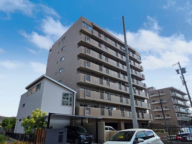 藤和シティコープ国府宮Ⅱの外観写真。隣接する戸建て住宅と並び、駐車場や車が写る全体像。青空と白い雲が背景に映えている。