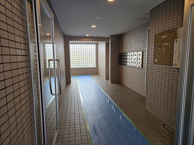 藤和シティコープ国府宮Ⅱのエントランスホール。床には養生シートが敷かれ、右側には集合ポストと掲示板、正面に大きなガラスブロック窓がある。