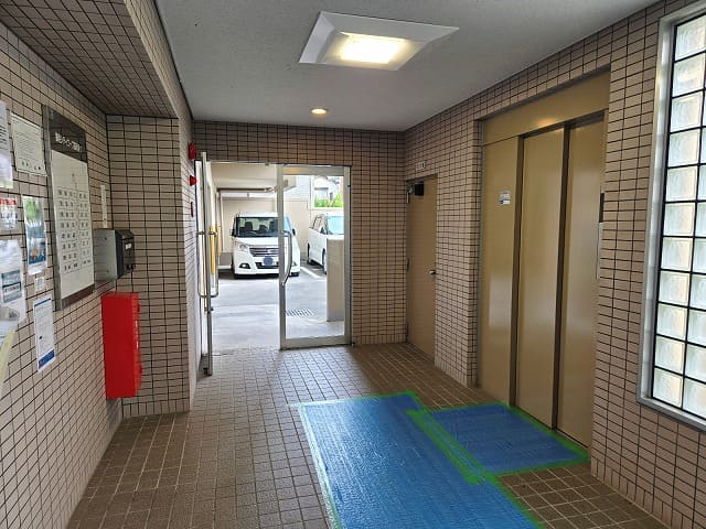 藤和シティコープ国府宮Ⅱのエレベーターホール。タイル張りの共用部にエレベーターが設置され、正面にはガラス扉の出入口と駐車場が見える。