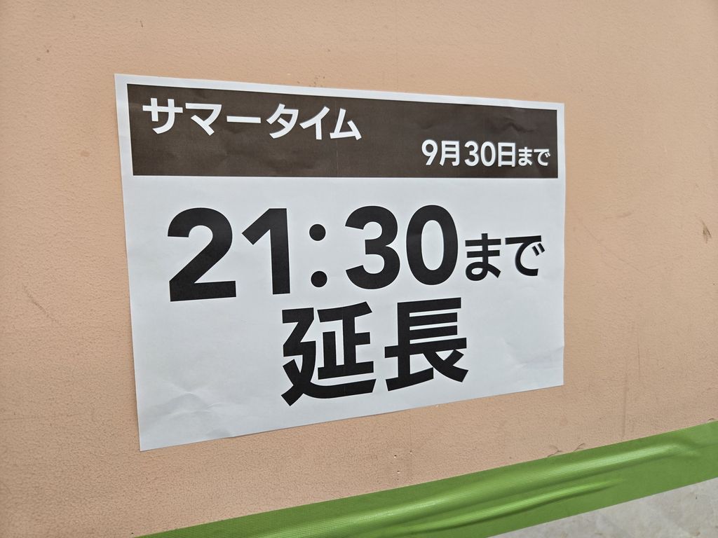 カネスエ国府宮店の外壁に貼られたお知らせの張り紙の写真。上部の黒い帯に「サマータイム 9月30日まで」とあり、その下に大きな黒文字で「21:30まで延長」と書かれている。壁はベージュ色で、下のほうに緑色のラインテープが見える。