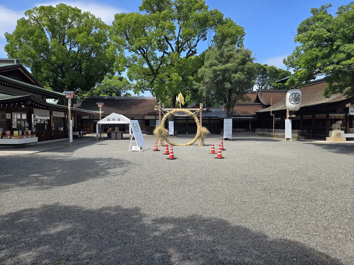 国府宮神社の拝殿前広場に設置された茅の輪を少し離れた場所から写した写真。晴れた青空の下、茅葺きの大きな輪の周りにオレンジ色のカラーコーンが並び、手前には国府宮の受付テントと案内看板が見える。奥には歴史ある社殿と大きな木々、赤い提灯が写っている。