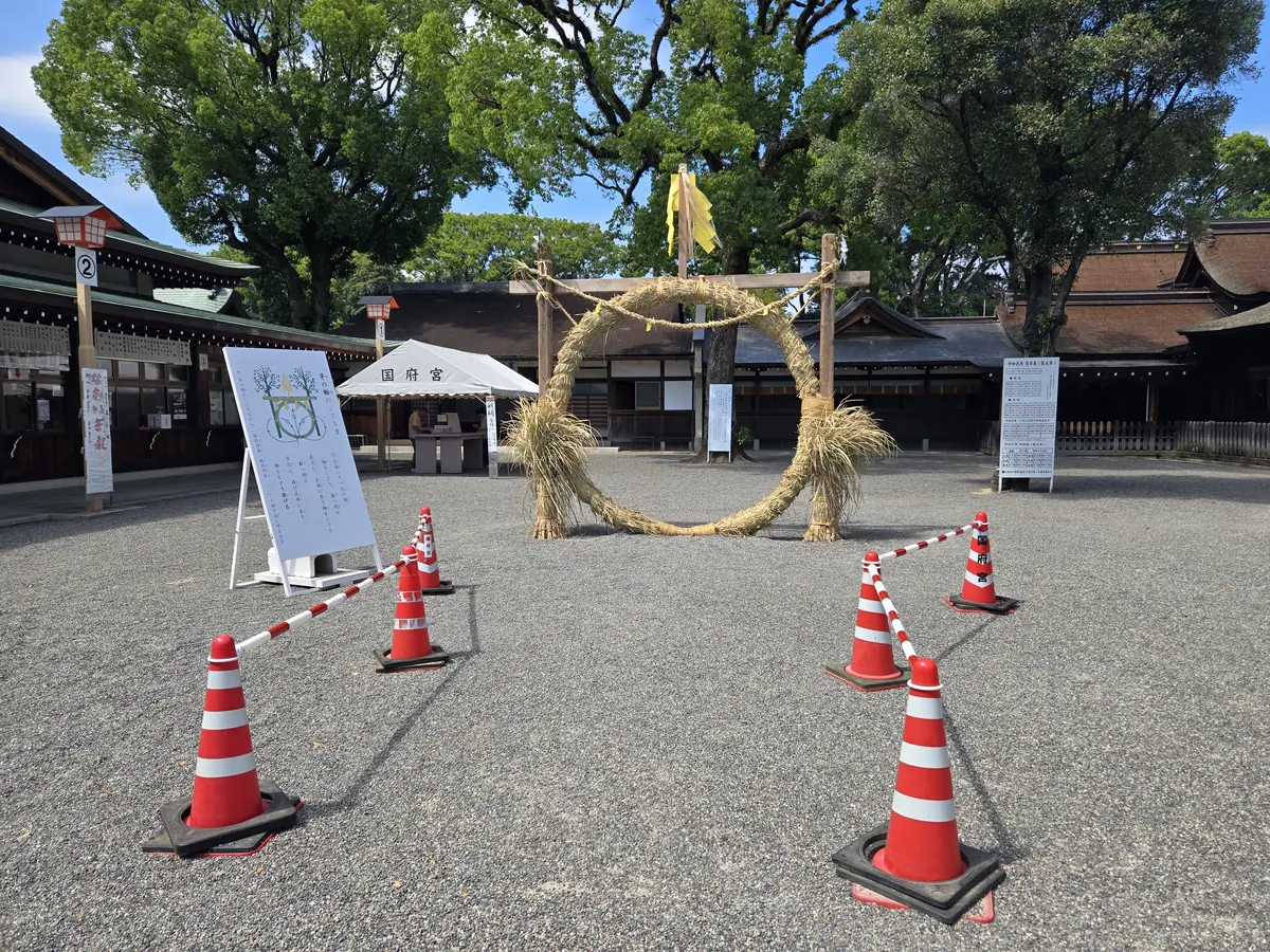 茅の輪を正面から写した写真。左手前に「茅の輪のくぐりかた」と書かれたイラスト入りの案内看板が置かれ、輪の周りをオレンジと白のカラーコーンとロープで囲ってある。背景には社殿と大きな木々、風にはためく黄色い旗が写っている。