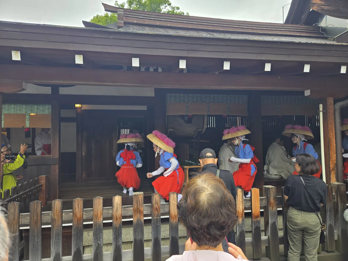 2026年6月28日の国府宮神社御田植祭で、本殿前に並び田植歌に合わせて舞う早乙女たち。菅笠の花飾り、青い上衣と赤い袴、後方の神職、手前で見守る参列者が写っている。